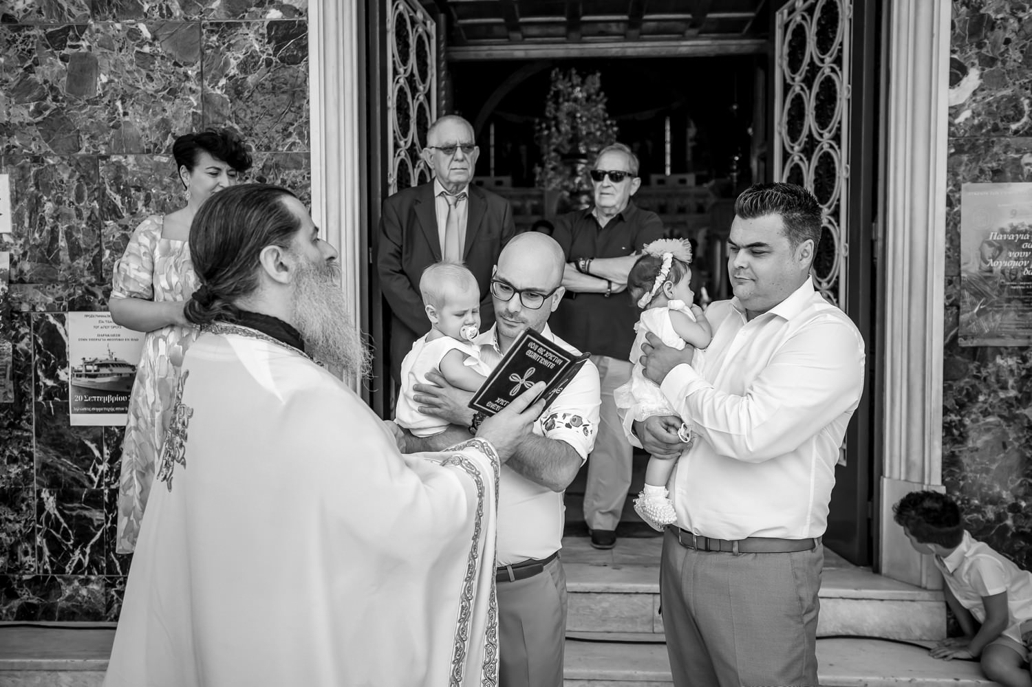 Φωτογράφηση βάπτισης διδύμων στη Λάρισα. Ασπρόμαυρη φωτογραφία ιερέα με νονούς και δίδυμα στην είσοδο της εκκλησίας κατά τη διάρκεια της κατήχησης.