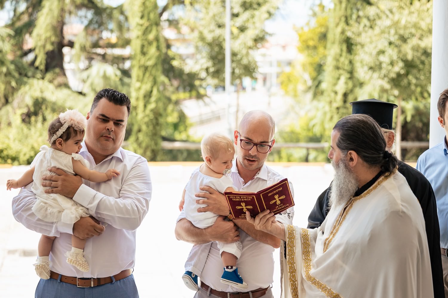 Φωτογράφηση βάπτισης διδύμων στη Λάρισα. Ιερέας διαβάζει κατηχητικές ευχές με νονούς που κρατούν τα δίδυμα έξω από την εκκλησία.