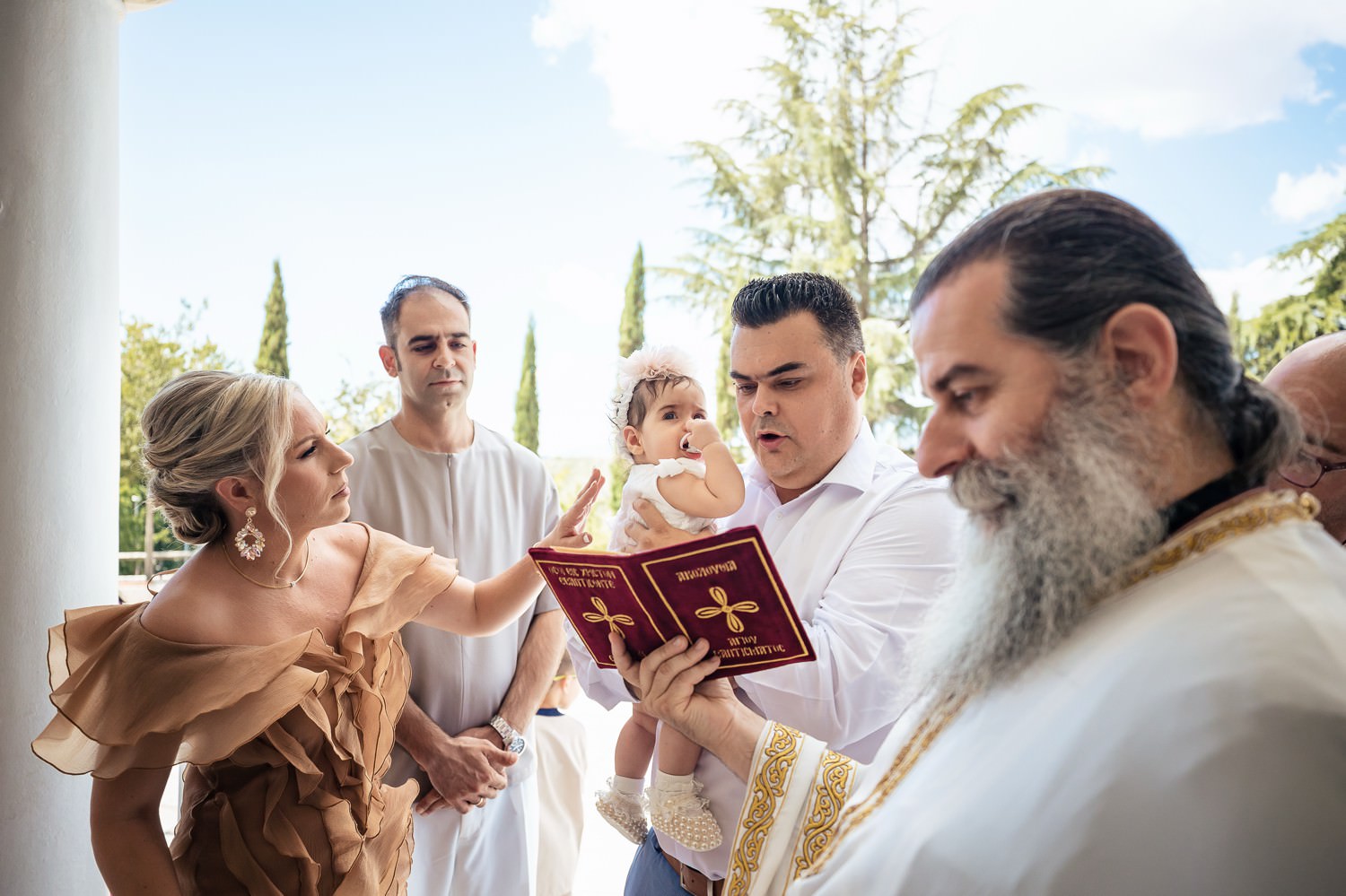 Φωτογράφηση βάπτισης διδύμων στη Λάρισα. Ιερέας διαβάζει κατηχητικές ευχές με γονείς και νονό που κρατά βρέφος έξω από την εκκλησία.